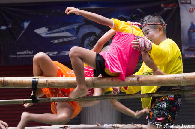 two kids boxing while balanced on a bamboo pole and one falling into water in Khao Phansa candle parade